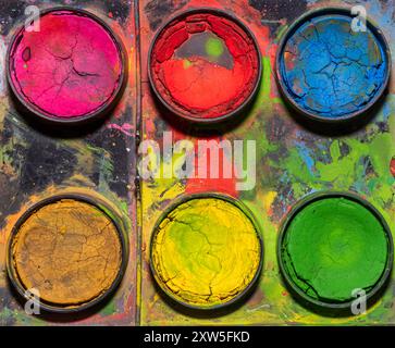 Immagine ravvicinata di una tavolozza di acquerelli ben utilizzata con sei pozzetti circolari riempiti di vernice secca e incrinata. I colori includono sfumature vivaci Foto Stock