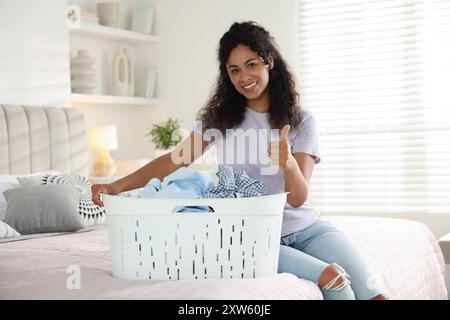 Donna felice con cesto pieno di biancheria che mostra i pollici sul letto a casa Foto Stock