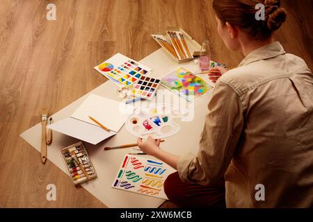 Donna che dipinge con acquerelli circondati da forniture d'arte su sfondo in legno Foto Stock