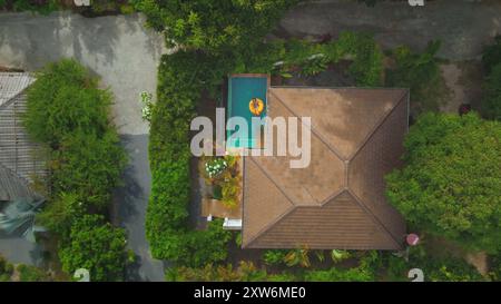 Vista aerea di una donna che si gode la sua vacanza galleggiando su un anello gonfiabile giallo in una piscina in una lussuosa villa tropicale Foto Stock