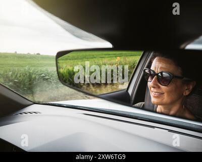 Faccia di una donna sorridente riflessa nello specchietto retrovisore mentre guidi un'auto e ti godi la vista panoramica della campagna. Foto Stock