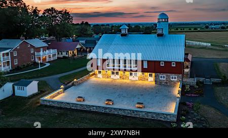 Ronks, Pennsylvania, USA, 19 luglio 2024 - tranquilla fattoria al tramonto con Un grande fienile rosso con Un Silo bianco, un cortile in pietra illuminato, Una vicina Brick House e ampi campi verdi sotto Uno splendido cielo al tramonto, creando Un ambiente tranquillo e pittoresco. Foto Stock