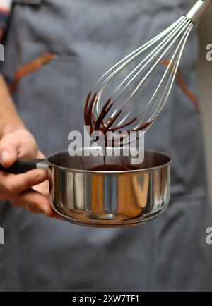 Chef che prepara una torta al cioccolato, primo piano delle mani che reggono una pentola. Cioccolato fuso su una frusta. Foto Stock