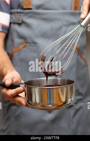 Chef che prepara una torta al cioccolato, primo piano delle mani che reggono una pentola. Cioccolato fuso su una frusta. Foto Stock