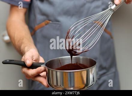 Chef che prepara una torta al cioccolato, primo piano delle mani che reggono una pentola. Cioccolato fuso su una frusta. Foto Stock