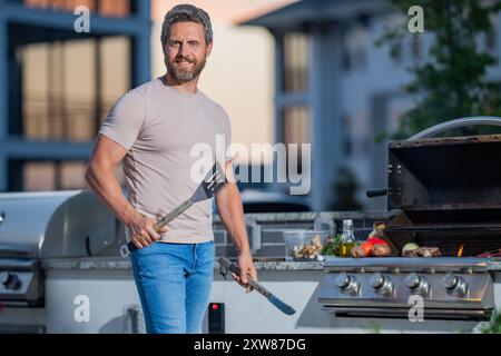 Uomo alla griglia del barbecue. Cuoco maschile preparazione barbecue all'aperto. Barbecue di carne, grill per picnic. Manzo arrosto. Cuoco preparare barbecue nel cortile della casa. B Foto Stock