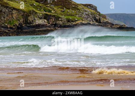 Le onde si infrangono sulla spiaggia sabbiosa, incontrando aspre scogliere sotto un cielo grigio, forti venti soffiano gli spruzzi Foto Stock