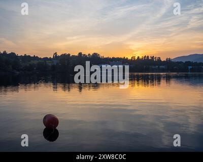 Un tranquillo tramonto dipinge il lago Placid in tonalità calde, con una boa che galleggia in primo piano e le maestose montagne Adirondack che si innalzano attraverso lo specchio L. Foto Stock
