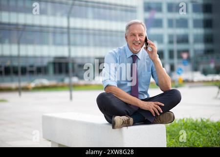 Ritratto di un uomo d'affari sorridente che usa il suo smartphone mobile all'aperto mentre è seduto su una panchina in città Foto Stock