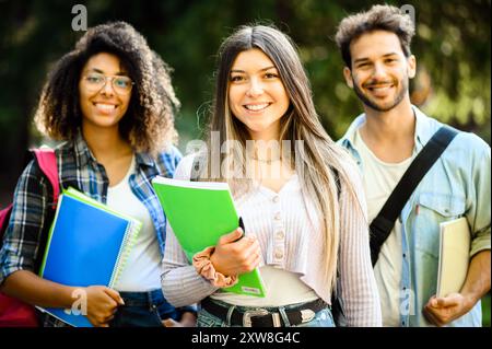 Tre studenti universitari felici che si trovano fuori dal campus, tengono libri e si preparano per le lezioni, emettono emozioni e cameratismo mentre si preparano Foto Stock