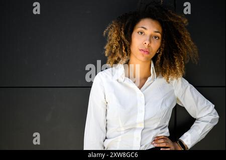 Una giovane donna professionale con i capelli ricci si trova di fronte a una moderna parete nera, indossa una camicia bianca e posa con la mano sull'anca Foto Stock