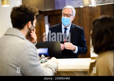 Il responsabile senior dell'hotel in una maschera parla con gli ospiti alla reception, concentrandosi sulla sicurezza e l'igiene durante la pandemia Foto Stock