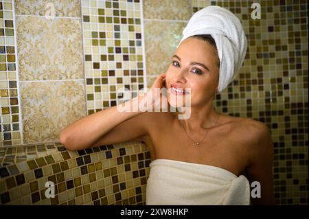Una bella donna sorride mentre ti godi un rilassante bagno turco in un bagno turco tradizionale. Sta indossando un asciugamano sulla testa e un altro intorno a lei Foto Stock