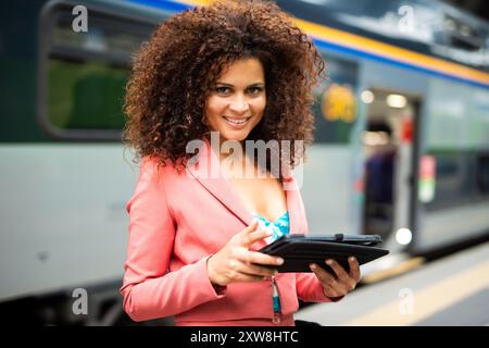 Un'elegante donna d'affari aspetta con fiducia in una stazione ferroviaria affollata, utilizzando un tablet digitale con un sorriso felice, che unisce la tecnologia al suo compagno di viaggio Foto Stock