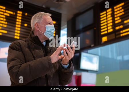 Uomo d'affari mascherato che utilizza il suo smartphone mentre attende il concetto di mobilità in treno, covid e coronavirus Foto Stock