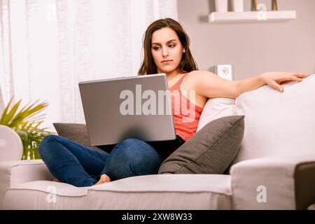 La donna si rilassa sul divano di casa, navigando su Internet con il suo laptop. È concentrata, si connette con il mondo online in un'immagine di stile di vita moderno Foto Stock