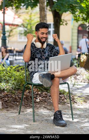 Giovane indiano che usa un computer portatile per celebrare la vittoria di buone notizie sui messaggi, la vittoria del jackpot della lotteria, giveaway online all'aperto. Il giovane turista felice si siede nel parco urbano della strada. Stili di vita della città. Foto Stock