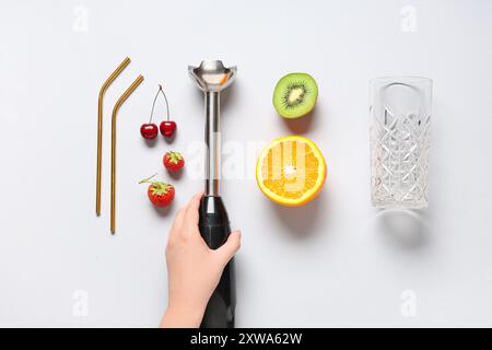 Donna che tiene in mano il frullatore a immersione e diversi ingredienti per preparare frullati freschi su sfondo bianco Foto Stock