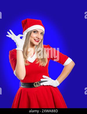 La giovane bionda Babbo Natale sorride alla macchina fotografica e fa un buon segno con la mano su sfondo blu Foto Stock