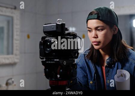 Ritratto di una giovane donna asiatica che utilizza apparecchiature fotografiche di fascia alta in ambienti interni, concentrandosi sulla produzione di contenuti visivi con dedizione e precisione Foto Stock