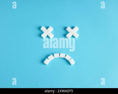 Consumo eccessivo di zucchero. Pericoli di un eccessivo apporto di zucchero. Rischio di diabete e problemi di salute. Faccia sorridente di emoticon morta fatta di cubetti di zucchero su blu Foto Stock