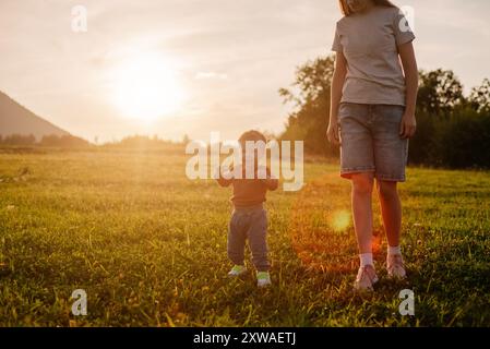 Il bambino carino va dalla sua giovane madre nel parco estivo impara a camminare per primi passi. I primi passi nella natura. Mamma insegna al figlio a camminare sullo sfondo Foto Stock