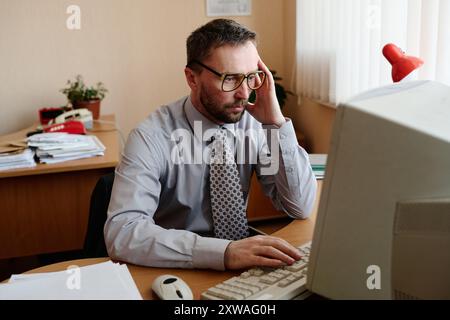 Ritratto di un uomo caucasico seduto alla scrivania, che utilizza un computer in un ufficio, indossa gli occhiali e tiene la testa premurosa Foto Stock