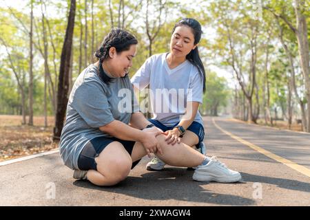 Donna in sovrappeso con lesioni al ginocchio seduta sulla pista da corsa in un parco di corsa tenendo il ginocchio doloroso mentre la sua amica di corsa seduta besi Foto Stock