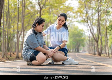 Donna in sovrappeso con lesioni al ginocchio seduta sulla pista da corsa in un parco di corsa tenendo il ginocchio doloroso mentre la sua amica di corsa seduta besi Foto Stock
