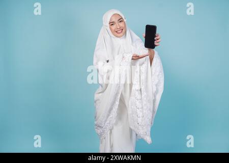 Bella giovane donna musulmana asiatica in abito da preghiera che presenta uno spazio di copia sullo schermo del suo smartphone, isolato da sfondo blu. Concetto Hajj. Foto Stock