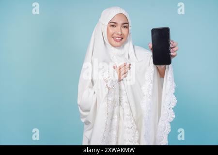 Bella giovane donna musulmana asiatica in abito da preghiera che presenta uno spazio di copia sullo schermo del suo smartphone, isolato da sfondo blu. Concetto Hajj. Foto Stock