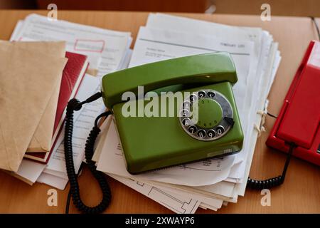 Primo piano di un telefono con quadrante rotativo verde d'epoca sopra una pila di documenti e file su una scrivania di legno, con un telefono rosso nelle vicinanze, che rappresenta l'allestimento di un ufficio retrò Foto Stock