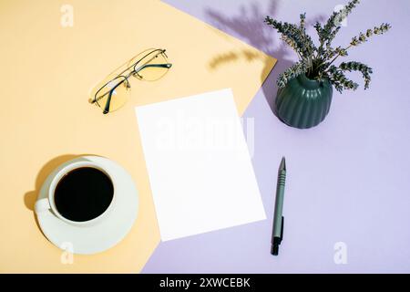 Scrivania con carta bianca, bicchieri, tazza da caffè e fiocchi lavanda in vaso su sfondo beige e viola bicolore alla luce del sole. Vista dall'alto, mockup. Foto Stock