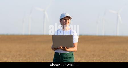 Donna agricoltrice che indossa un berretto bianco e una t-shirt con laptop sul campo agricolo al tramonto. Turbine eoliche sullo sfondo. Foto Stock