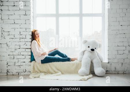 ragazza incinta dai capelli rossi in una blusa leggera e jeans blu con un orsacchiotto sulla finestra Foto Stock