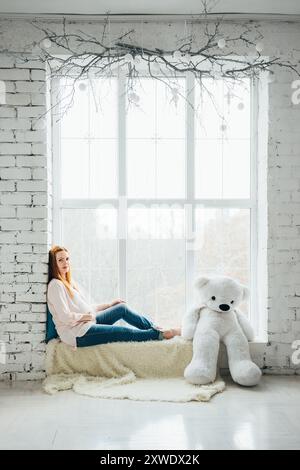 ragazza incinta dai capelli rossi in una blusa leggera e jeans blu con un orsacchiotto sulla finestra Foto Stock