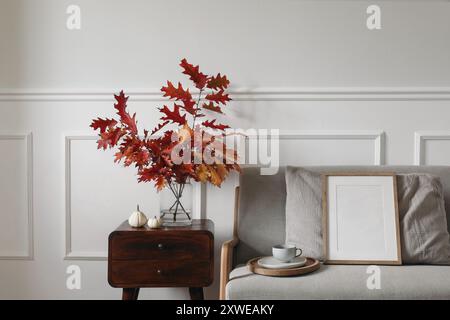 Foglie di quercia rossa, rami in vaso su comodino in legno. Divano in lino della metà del secolo con tazza di caffè. Mockup con cornice in legno vuota. Un po' bianco Foto Stock