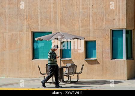 Lo smartphone toting Student Walks passa davanti al tavolo e alle sedie coperti da ombrelloni del patio dell'Università del New Mexico, Albuquerque, NEW MEXICO, aprile 2024 Foto Stock