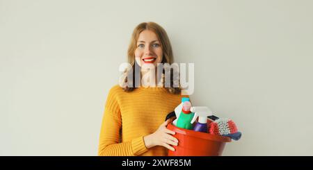 Faccende domestiche, giovane donna sorridente e felice tiene in mano un secchio di plastica di prodotti per la pulizia e detergenti per pulire la casa Foto Stock