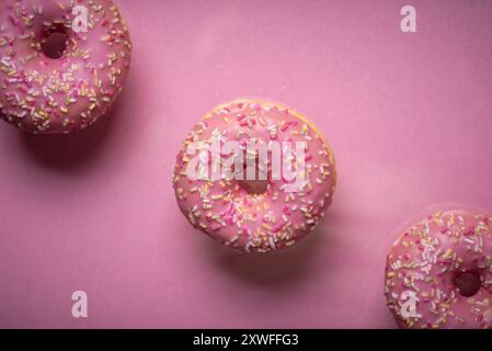 Un'immagine ravvicinata di tre ciambelle rosa vivaci ricoperte di colorate macchie, su uno sfondo rosa brillante, che catturano l'essenza del dolce e viziato Foto Stock