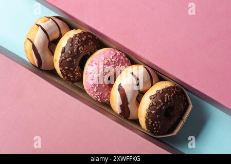 Una disposizione visivamente attraente di sei ciambelle con un mix di cioccolato e glassa rosa, disposte ad arte in un vassoio di cartone, adagiato su un rosa a contrasto Foto Stock