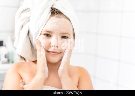 Ritratto di una bella bambina pre-adolescente davanti a uno specchio in un bagno bianco con un asciugamano sulla testa, si lava il viso con sapone o con il suo Foto Stock