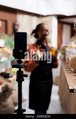 Primo piano di uno smartphone e un cavalletto utilizzati da una donna nera per promuovere prodotti biologici locali. Una donna afroamericana negoziante che pubblicizza opzioni fresche e salutari. Concentrati sul primo piano. Foto Stock