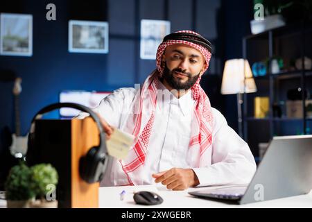 Bell'uomo d'affari che indossa un abito islamico, seduto nel suo ufficio a lavorare sul suo portatile e a scrivere appunti. Giovane musulmano che mette la sua nota adesiva su una tavola di legno con le cuffie. Foto Stock