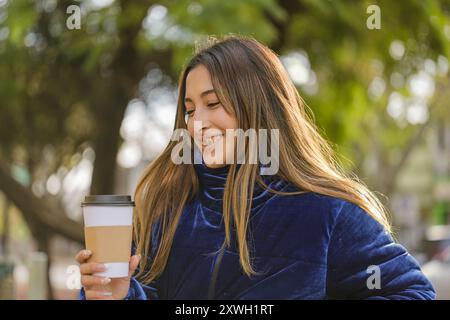 Sorridente ragazza latina che beve caffè su una panchina del parco pubblico. Foto Stock