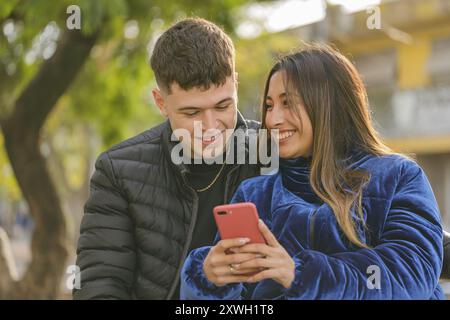 Sorridente ragazza latina che mostra il suo cellulare a un ragazzo caucasico in un parco pubblico. Foto Stock