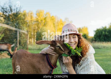 Donna sorridente in un cappello che si lega a una capra in un ambiente rurale. Gioiosa donna di mezza età che offre partenze a una capra in una giornata di sole Foto Stock