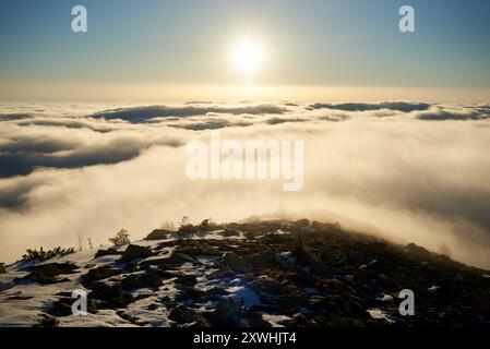 Alba sulla cima innevata della montagna. La calda luce dorata del sole che sorge bagna le nuvole in delicate sfumature di arancione e rosa. Neve e rocce sparse in primo piano aggiungono texture e profondità alla scena. Foto Stock