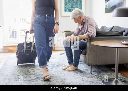 Symbolfoto zum Thema Trennung in der Partnerschaft. Eine Frau zieht einen Rollkoffer durch ein Wohnzimmer waehrend ihr Mann deprimiert auf dem Sofa si Foto Stock