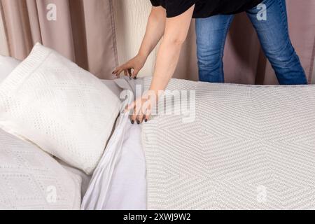 Una donna dispone meticolosamente una coperta bianca su un letto, soffici cuscini per creare un ambiente accogliente. Il letto non fatto presenta lenzuola leggermente spiegazzate, una Foto Stock